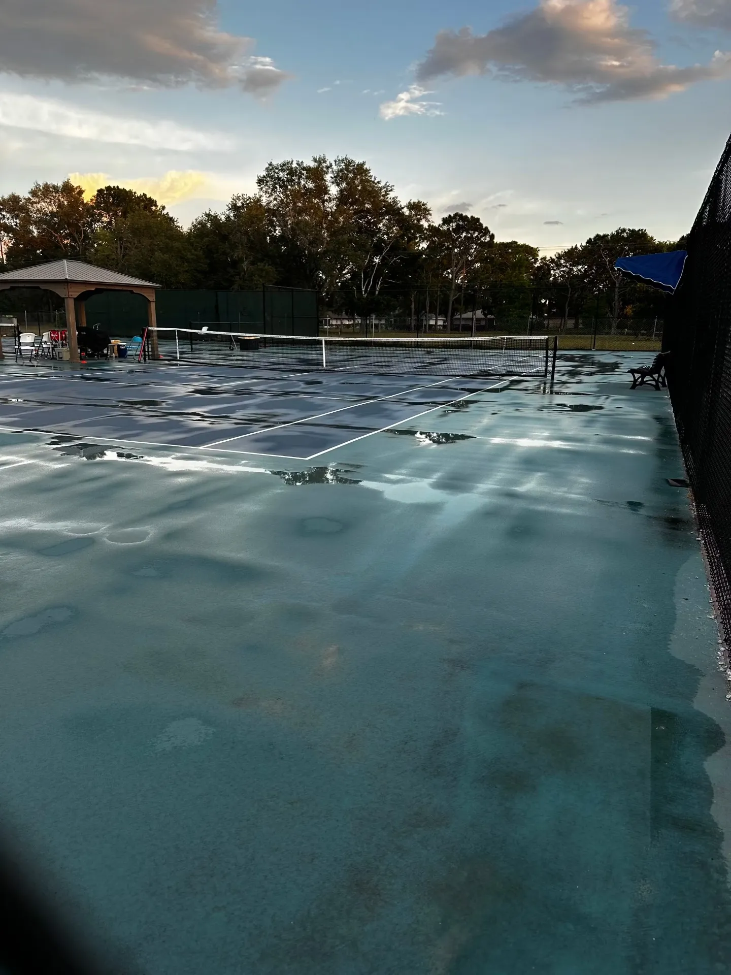 Standing water across a blue and green tennis court beside a pavilion.